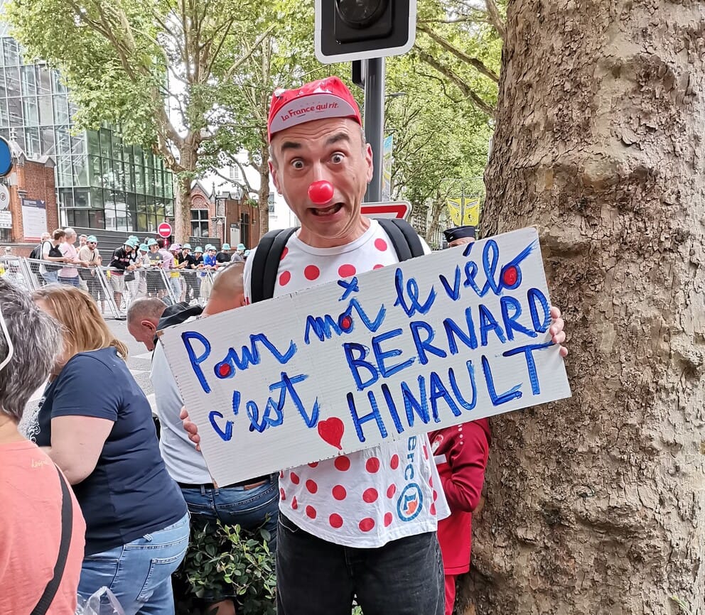 Un homme grimé en clown avec un nez rouge, une casquette "La France qui rit" et un maillot à pois du Tour de France tient une pancarte peinte à la main où l'on peut lire : "Pour moi le vélo c’est Bernard Hinault", écrit en bleu et rouge. Il se tient debout au bord de la route, au départ du Tour de France 2025 à Lille.